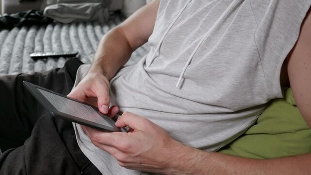 A young man holds a thin electronic e-reader device while sitting comfortably on a grey textured bed. He uses his thumbs to scroll through pages of text in a relaxed indoor setting.