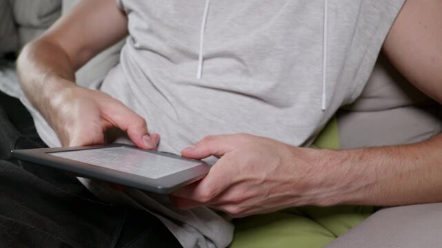 A man sits on a comfortable sofa while reading text on a grey e-book reader tablet. His hands gently hold the device as he scrolls through the digital pages in a cozy indoor setting.