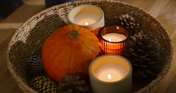Camera is closing in as flames are flickering, casting warm glow on table pumpkin, gold pinecone