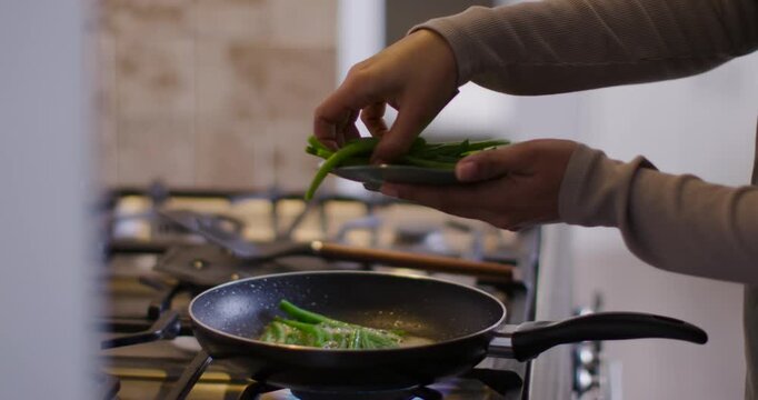 Hands are placing plate-held green beans flat into sizzling pan over blue gas flame to cook
