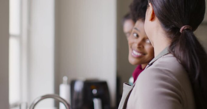 Diverse women friends talking laughing reaching tap and washing dishes at kitchen sink in aprons