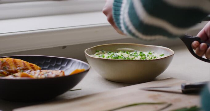 Woman in striped sweater bringing scissors, snipping chives over bowl, garnishing dish on counter
