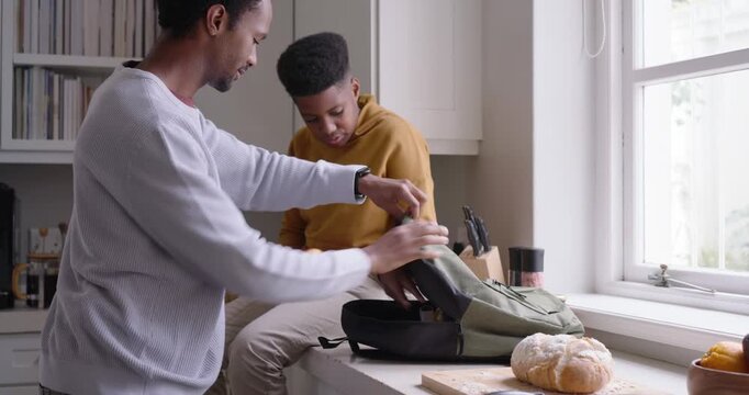 Father and teenage son packing backpack in kitchen after dad bringing lunchbox, hugging for school