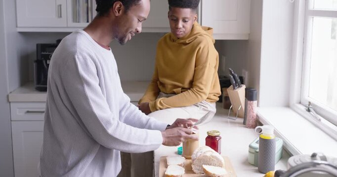 African American dad at counter holding peanut butter jar to teen, spreading on bread making snack