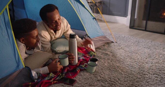 African American father and teen son smiling, pouring thermos into mugs in blue tent, sharing drink