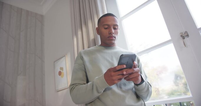 African American man standing near paned window, camera pulling back, reading, scrolling smartphone