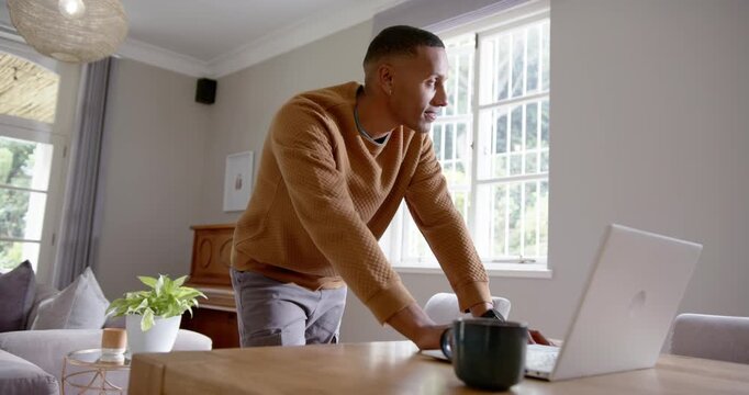 African man bending leaning over table using laptop touching mug brown knit working at home