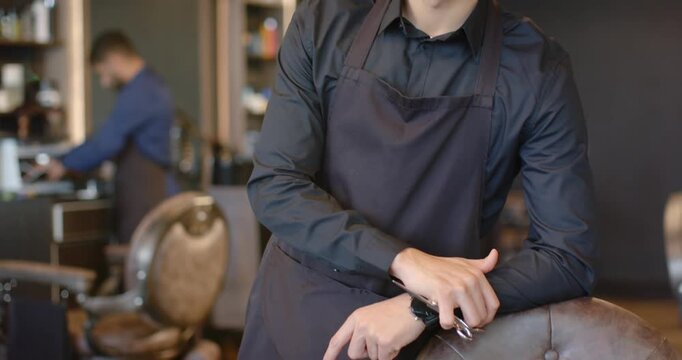 Male barbers, camera pulling back, barber leaning on chair holding scissors, preparing cut in shop