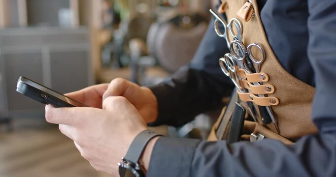 African man barber picking up phone and checking messages, leather apron and scissors in salon
