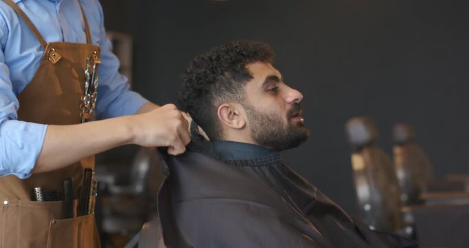 Male barber in apron pulling cape around male client neck in shop, securing strap for haircut