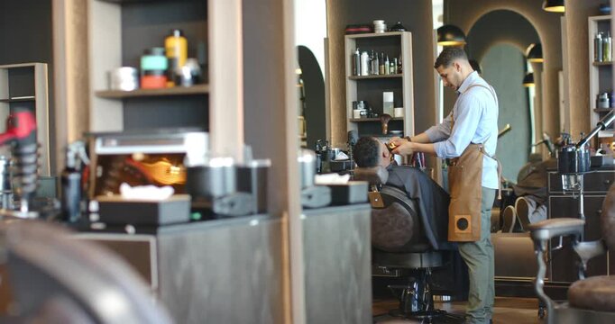 Male barber trimming male client after adjusting tools at salon using clippers and comb for haircut