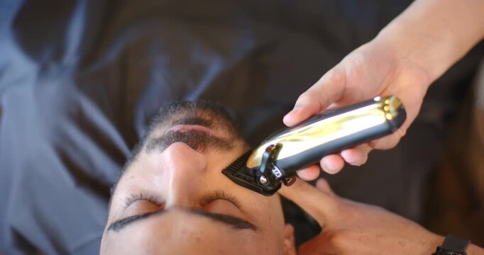 African male barber trimming male client facial hair, picking gold clipper and using comb on cape