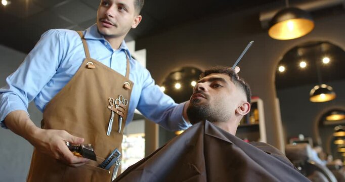 Barber trimming Middle Eastern client's beard at barbershop after placing comb, checking symmetry