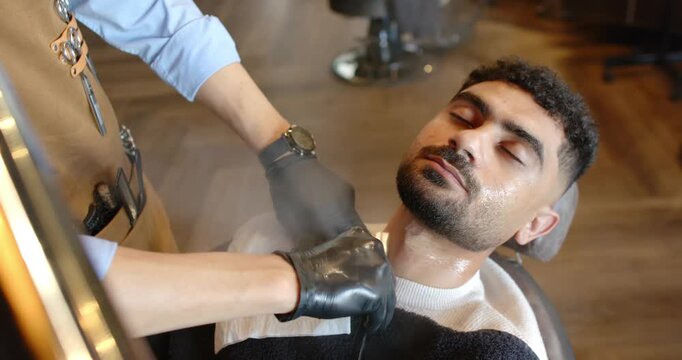 Client reclining in barber chair with bib, barber performing straight-razor shave for grooming