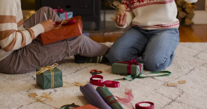 African American couple wearing Santa hats wrapping gifts on rug, tying twine by fire for holiday