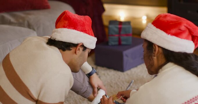 Couple in Santa hats preparing gifts at home, slipping sock on foot, writing tag, high-fiving