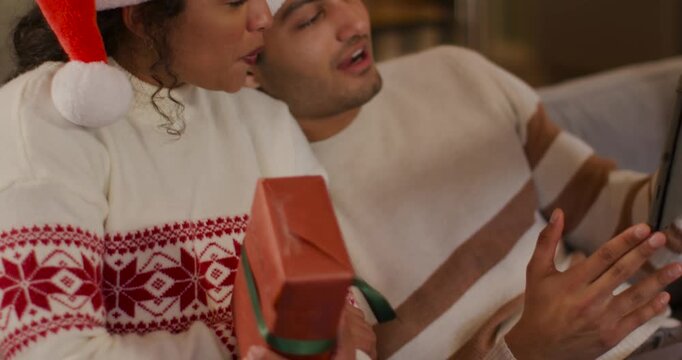 African American couple sitting on couch wearing Santa hats, holding holiday gift, gesturing tablet