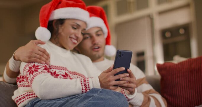 African American couple sitting on sofa in Santa hats, woman tapping phone, couple sharing clip