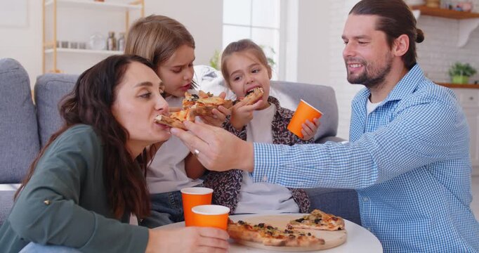 Happy young mother, father with two cute children eating slices of pizza and talking together sitting in the living room at home. Family leisure and delivery service concept. 4k video.