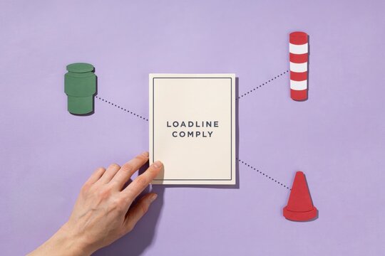 A hand holds a sign with loadline comply surrounded by traffic cone, green bucket, and red cylinder on purple background