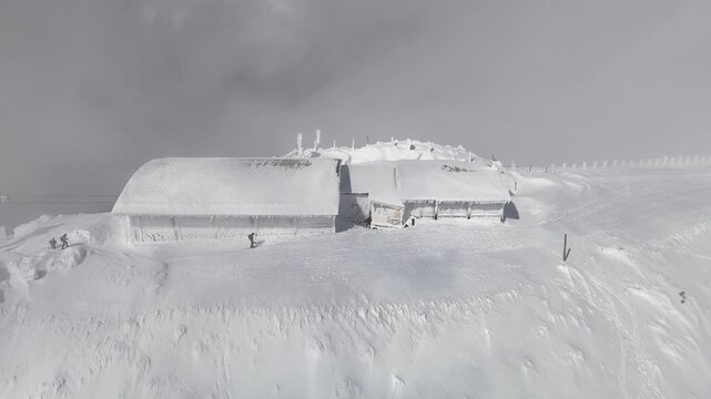 Aerial drone footage of the snow covered summit near Bola del Mundo at Puerto de Navacerrada, Sierra de Guadarrama, Madrid, Spain, with skiers and ice covered buildings