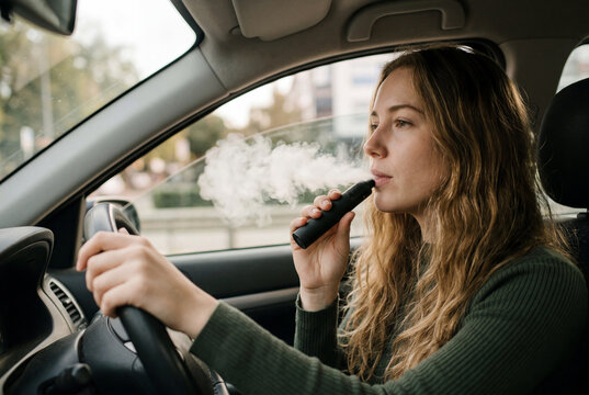 Young woman driving a car while exhaling thick smoke from an electronic cigarette device