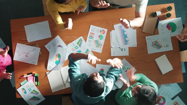 Top view of smart people throw paper and giving high five at meeting table with financial document and stock market analysis paper scattering around. Aerial view of team celebrate project. Symposium.