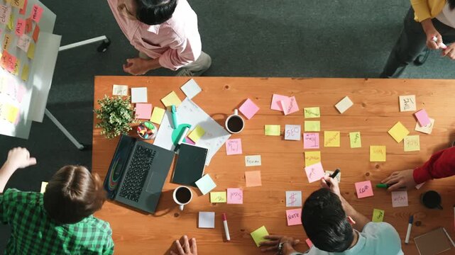 Top view of skilled business people discuss plan at meeting room while writing idea at sticky notes and stick at whiteboard. Group of diverse team working together to planning strategy. Convocation.