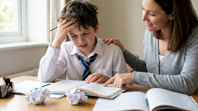 Schoolboy struggling with difficult homework assignments receives gentle academic support and guidance from his patient mother at the kitchen table.