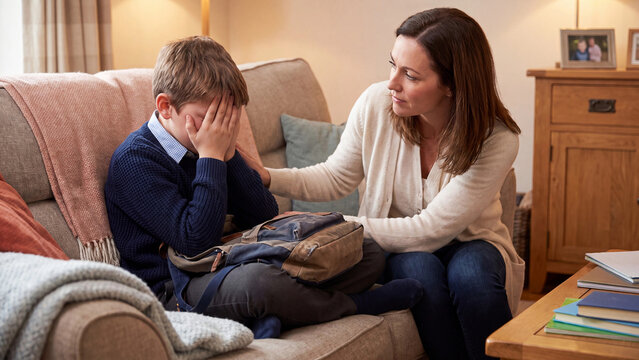 Mother provides emotional support to her young son who sits on the sofa with his head in his hands after returning home from a challenging day at school.