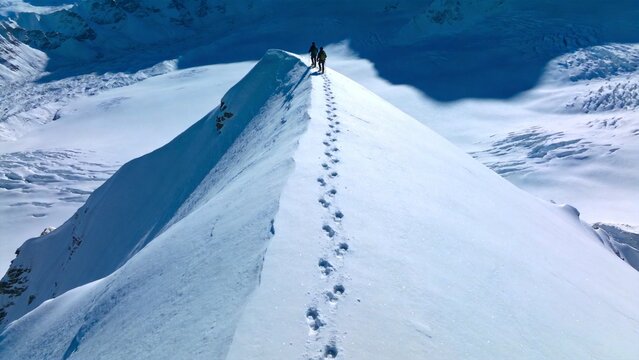 Summit Conquerors: Footprints trail a steep, snow-covered mountain ridge, leading two climbers towards a majestic summit under a clear, expansive sky.