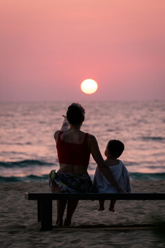 Mother and son sitting on a beach bench watching the glowing sunset over calm ocean