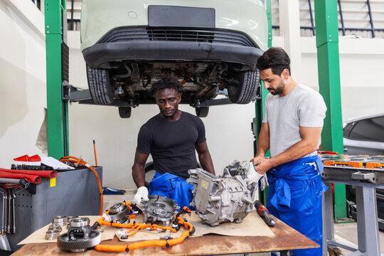 Professional EV technicians servicing an electric drive unit and high-voltage electrical components beneath a lifted electric vehicle in a modern workshop.