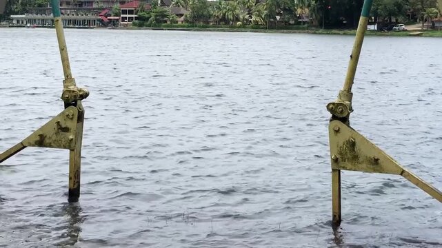 Weathered metal linkage stands partly submerged in rippling lake water, offering industrial, corrosion, maintenance, infrastructure, engineering, and waterfront concepts.