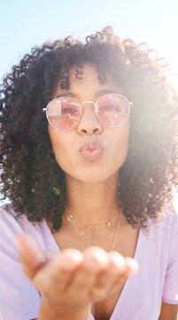 A young woman with curly hair wearing tinted sunglasses blows a kiss towards the camera in a bright outdoor setting with beautiful lens flare glow