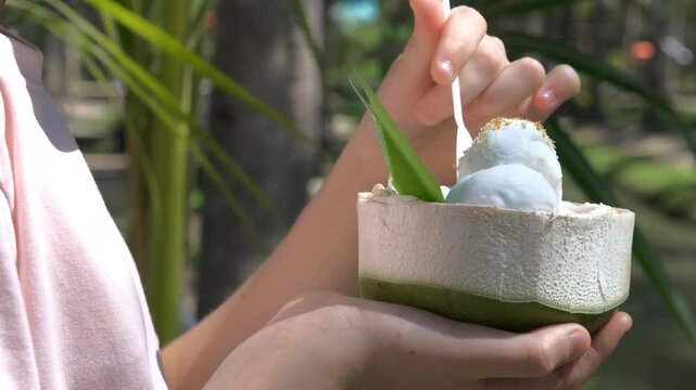Close-up of a teenage girl taking a bite of coconut ice cream with a spoon surrounded by greenery, slow motion. Tropical dessert concept