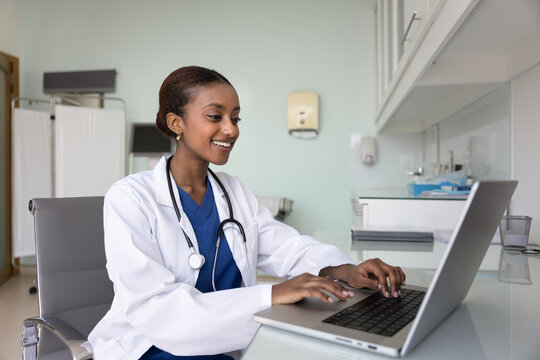 Happy young African general practitioner woman working at computer in medical office, typing report, records on laptop, smiling, chatting with patients, providing telehealth service