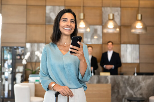 Hotel guest standing in lobby with suitcase, using mobile application on smartphone to check reservation details and booking confirmation before check-in. Travel planning, digital tourism services