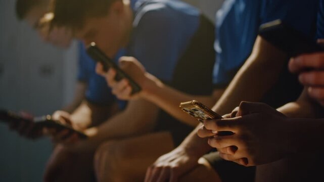 Young Male Football Players Relaxing on Benches in Sports Changing Room, Silently Using Smartphones. Concept of Modern Youth Digital Routine, Digital Isolation Among Gen Z Athletes, Youth Lifestyle.