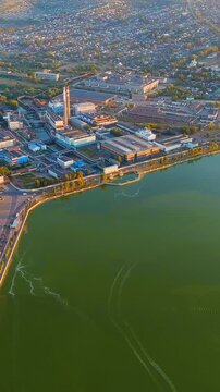 Vertical video. Russia, Votkinsk. Votkinsk Reservoir embankment, industrial waterfront with factories and dam infrastructure, aerial panoramic view at sunset with golden light over the water. Drone f