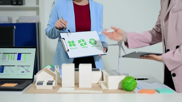 Two female professionals presenting an eco-friendly smart city project with solar houses, wind turbines, and a recycling plan.
