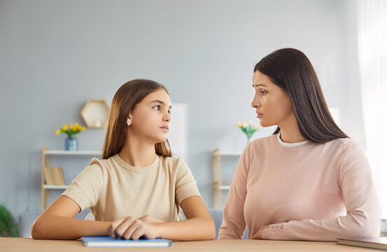 Serious mother and teenage daughter sitting at table, looking at each other with concern. Caring parent having deep conversation with child, discussing important matters, emotions or personal issues.