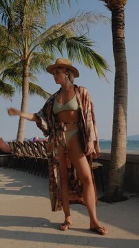 Stylish woman in straw hat and patterned kimono at tropical waterfront with colorful beach lounge.