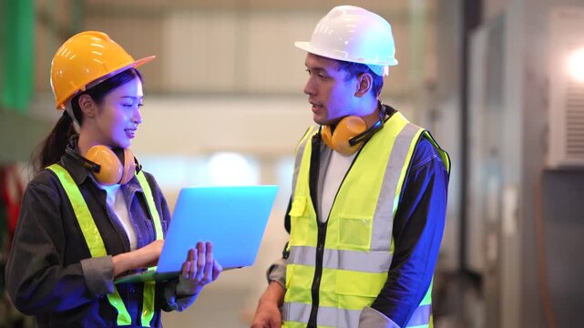 Male and female factory engineers walk through production area while discussing operations. Female engineer carries laptop and explains production details during engineering collaboration.