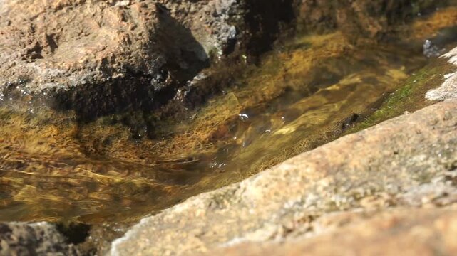the view of a small natural stream or water puddle flowing through cracked and weathered brown stone boulders, showcasing detailed geological textures.