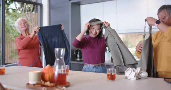 Diverse female friends plus senior finding aprons in kitchen, lifting straps and preparing meal