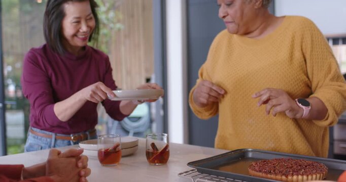 Diverse senior women in mustard, maroon tops approaching counter cutting pecan tart sharing dessert