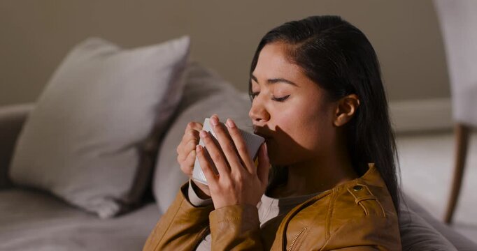 African American woman in brown jacket lifting white mug, inhaling aroma, sipping, tasting at home