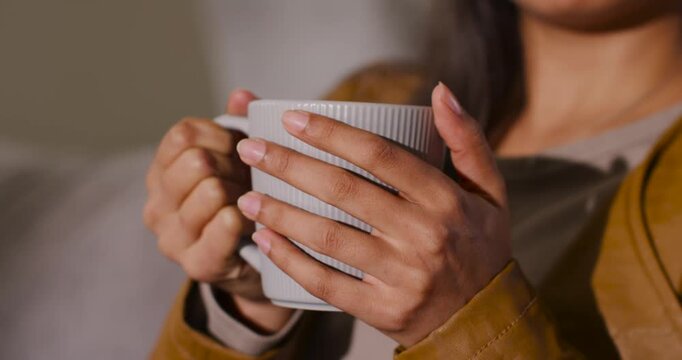 African American female cradling ribbed ceramic mug on sofa, wearing tan jacket seeking warmth