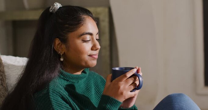 Indian woman cradling dark-blue mug on sofa at home, inhaling rising steam, sipping, seeking calm